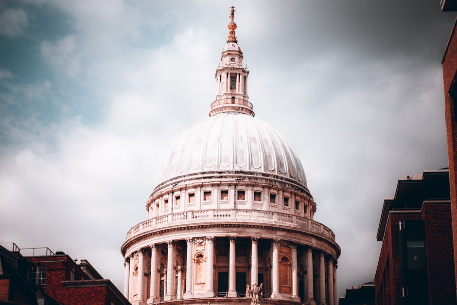 A detailed view of St. Paul's Cathedral in London, England, showing its iconic large white dome topped with a lantern and golden cross. The cathedral's classical architecture features columns and ornate detailing at the base of the dome. Surrounding the cathedral, modern brick buildings and structures are partially visible on either side of the image, with the overcast sky providing a neutral backdrop. This scene illustrates a prominent London landmark, captured during daytime, with natural lighting highlighting the intricate details of the dome and facade. The image is relevant to home relocation and furniture transport, as it reflects an area often involved in house removals within London, supported by professional removal services like Man with Van St Pauls Cray.