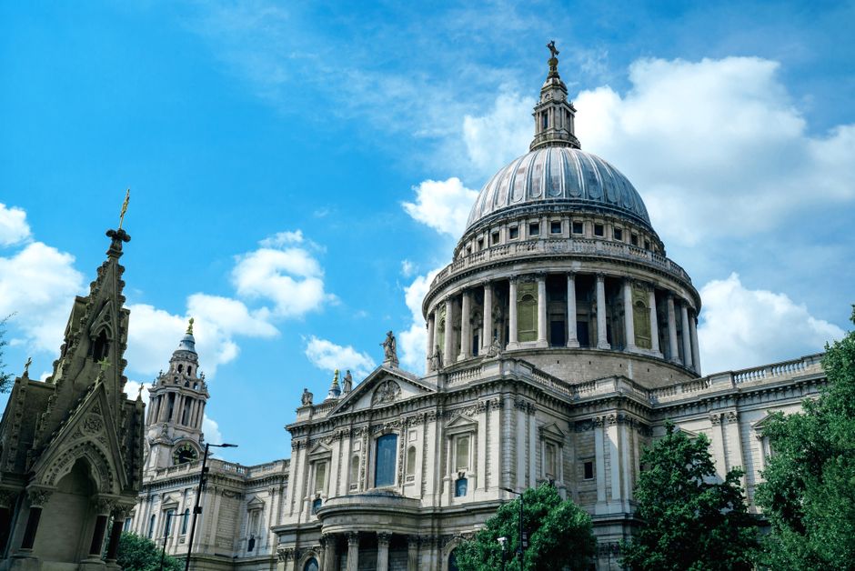 An exterior view of St. Paul's Cathedral in London, featuring its large central dome topped with a lantern and cross, surrounded by other historic church buildings with ornate gothic architecture, pointed arches, and decorative spires. The sky above is bright blue with scattered white clouds, and the cathedral is partially framed by green trees at the bottom right of the image. The scene captures the architectural details of the stone facade, including columns, statues, and intricate carvings, illustrating a prominent landmark in the context of a home relocation or moving process undertaken by a professional removals service such as Man with Van St Pauls Cray, which specialises in house removals and furniture transport within the UK.