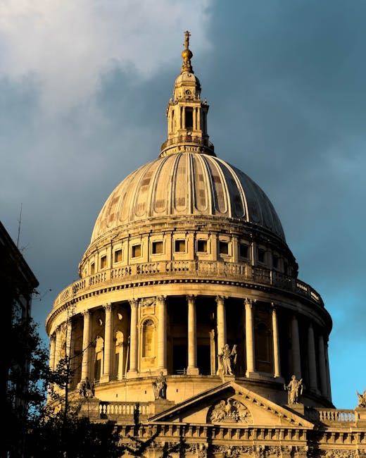 A detailed view of St. Paul's Cathedral in London, England, showing its iconic large white dome topped with a lantern and golden cross. The cathedral's classical architecture features columns and ornate detailing at the base of the dome. Surrounding the cathedral, modern brick buildings and structures are partially visible on either side of the image, with the overcast sky providing a neutral backdrop. This scene illustrates a prominent London landmark, captured during daytime, with natural lighting highlighting the intricate details of the dome and facade. The image is relevant to home relocation and furniture transport, as it reflects an area often involved in house removals within London, supported by professional removal services like Man with Van St Pauls Cray.