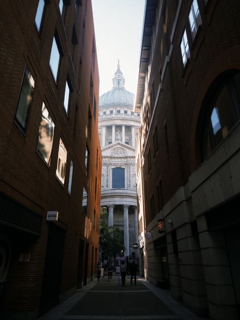 A narrow urban street framed by tall brick and concrete buildings on either side, leading towards St Paul's Cathedral with its iconic dome and columns visible in the background. The sky is partly cloudy, and a few pedestrians are walking along the pavement, suggesting a typical city environment. The scene captures the exterior setting relevant to house removals and moving logistics in central London, with the cathedral providing a recognizable landmark for the area. Man with Van St Pauls Cray may utilize similar city streets for residential or commercial furniture transport during home relocations or packing and moving services in the area.
