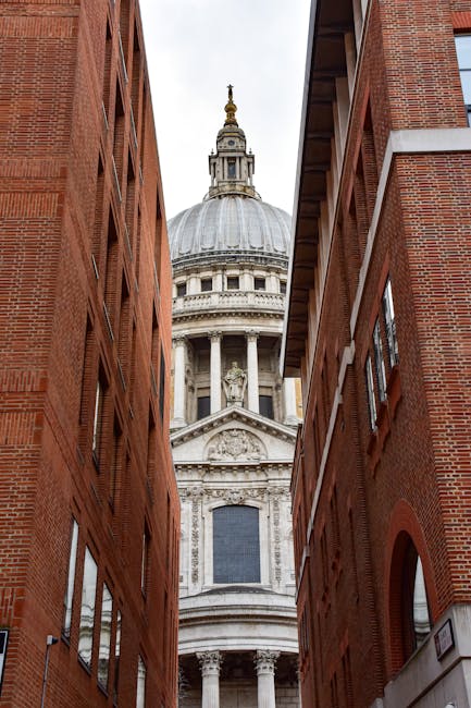A narrow alleyway between two red-brick buildings leading to St. Paul's Cathedral with its large, domed roof and ornate architectural details visible in the background, set against an overcast sky. The image captures the urban environment typical of a city centre, highlighting the street’s tight space and the historic cathedral’s prominence, suitable for illustrating city relocation or house removal services near central London or similar urban settings, as provided by Man with Van St Pauls Cray.