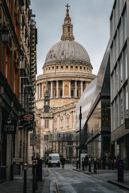 A narrow city street lined with modern glass-fronted buildings on the right and historic, ornate buildings on the left, leading towards the prominent dome of St. Paul's Cathedral in London. In the foreground, there are a few pedestrians walking, and a white van is parked near the curb, with some people engaged in loading or unloading items. The street surface appears wet, possibly after rain, and the sky is overcast. This urban scene captures the typical environment for home relocation or furniture transport services in central London, with the cathedral serving as a recognizable landmark. The image suggests a busy area suitable for house removals, showing the logistical setting for loading and unloading household items close to historic and modern architecture. Man with Van St Pauls Cray will coordinate moves within such city environments, involving careful navigation of parking zones and urban logistics for household or office relocations.