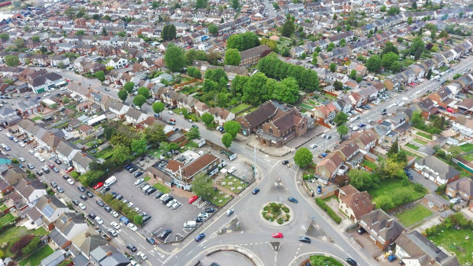 Aerial view of a residential neighbourhood in St Paul's Cray showing a roundabout at the intersection of several streets. Surrounding the roundabout are detached and semi-detached houses with tiled roofs, organized along tree-lined streets. There is a church or community building with a pitched roof near the intersection, surrounded by parking areas filled with cars. Green spaces with lush trees are visible throughout the area. In the foreground, part of a property with a driveway and parked vehicles can be seen, indicating proximity to home relocation and moving service areas. The overall scene captures a typical suburban environment, highlighting the residential layout and street infrastructure relevant to house removals and transport logistics, as managed by Man with Van St Pauls Cray.