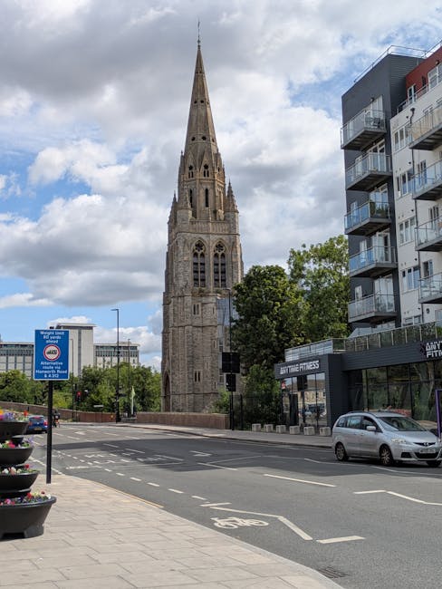 A street scene featuring a historic stone church tower with pointed spire and Gothic windows in the background, alongside modern multi-storey residential buildings with balconies on the right side. The road has white markings, including a bicycle lane, and cars are parked along the curb and driving past. In the foreground, there are planters with flowers on the sidewalk and a blue street sign indicating parking regulations. The scene is illuminated by daylight with partly cloudy skies overhead, providing clear visibility of the urban environment. This setting illustrates an area in St Paul's Cray suitable for residential relocations, and such visuals are relevant for the context of house removals and furniture transport services by Man with Van St Pauls Cray, supporting seamless home relocation procedures involving loading and unloading processes.