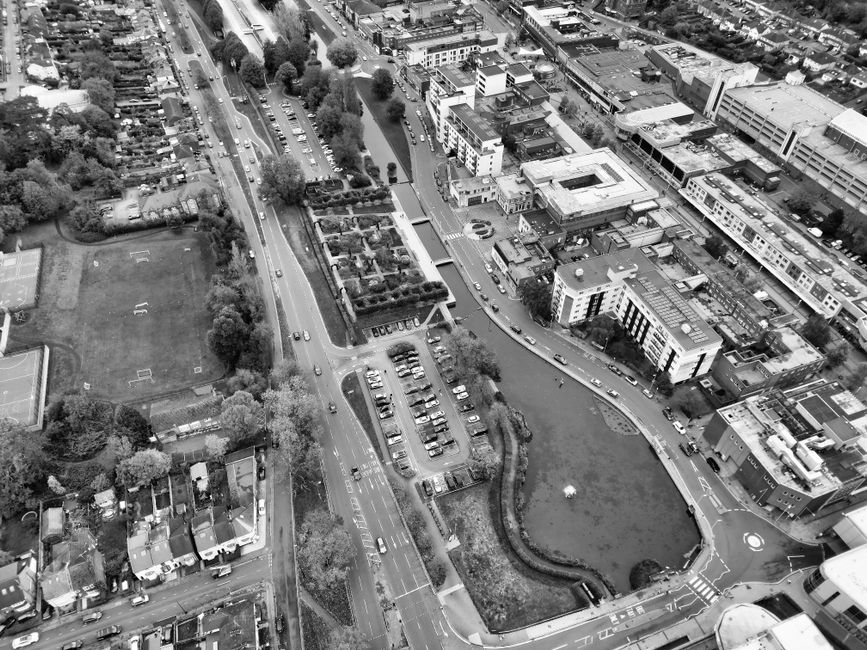 Aerial view of a residential neighbourhood in St Paul's Cray showing a roundabout at the intersection of several streets. Surrounding the roundabout are detached and semi-detached houses with tiled roofs, organized along tree-lined streets. There is a church or community building with a pitched roof near the intersection, surrounded by parking areas filled with cars. Green spaces with lush trees are visible throughout the area. In the foreground, part of a property with a driveway and parked vehicles can be seen, indicating proximity to home relocation and moving service areas. The overall scene captures a typical suburban environment, highlighting the residential layout and street infrastructure relevant to house removals and transport logistics, as managed by Man with Van St Pauls Cray.