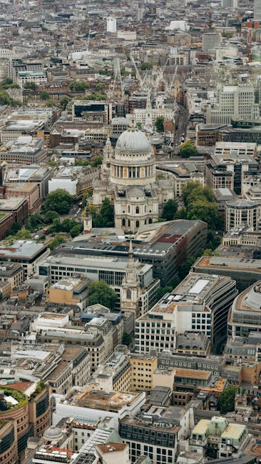 An aerial view of central London showing a cluster of urban buildings, streets, and green spaces, with St. Paul's Cathedral prominently positioned in the middle distance. The cathedral's large dome and classical architecture are clearly visible, surrounded by a mix of modern and historic structures, some with flat rooftops and others with pitched roofs. The scene includes a busy cityscape with closely packed office buildings, residential apartments, and commercial premises, illustrating typical city logistics. This image highlights the dense environment where local house removals and furniture transport services by Man with Van St Pauls Cray may operate, especially during home relocation processes involving loading, packing, and transport of household items in an urban setting.