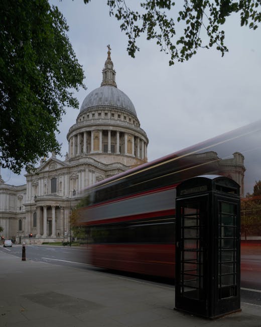 A black traditional British telephone box is positioned on the pavement in front of the historic St Paul's Cathedral in London, with its iconic dome and classical architecture visible in the background. The scene is set during overcast weather, and a red double-decker bus is passing briskly in front of the cathedral, creating a blurred motion effect that indicates movement. Green foliage from trees frames the top of the image, and the scene captures the busy city environment typical of London, highlighting elements relevant to urban moving services such as transport infrastructure and city landmarks. This setting reflects the context of house removals and relocation logistics, showcasing street-based transport and architectural features associated with city-wide moving and packing procedures, as would be managed by [COMPANY_NAME] for home relocation projects under the service category of removals.