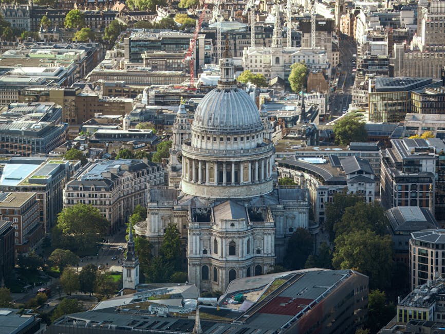 An aerial view of central London showing a cluster of urban buildings, streets, and green spaces, with St. Paul's Cathedral prominently positioned in the middle distance. The cathedral's large dome and classical architecture are clearly visible, surrounded by a mix of modern and historic structures, some with flat rooftops and others with pitched roofs. The scene includes a busy cityscape with closely packed office buildings, residential apartments, and commercial premises, illustrating typical city logistics. This image highlights the dense environment where local house removals and furniture transport services by Man with Van St Pauls Cray may operate, especially during home relocation processes involving loading, packing, and transport of household items in an urban setting.