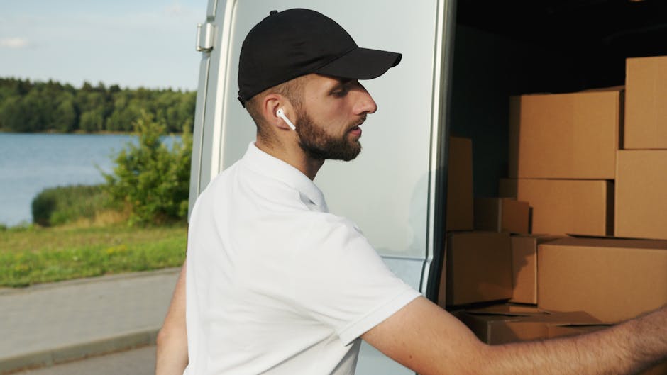 A man wearing a white t-shirt, dark trousers, and a baseball cap is walking beside a white commercial van parked on the street outside a residential property. The van is positioned parallel to the cur