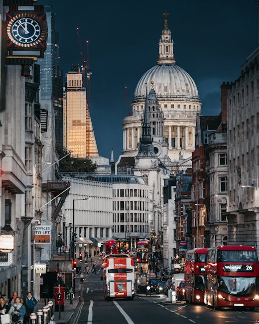 A busy city street scene in London during the evening with traffic including red double-decker buses and cars, leading towards the prominent St. Paul's Cathedral with its large domed roof and classical architecture illuminated against a dark blue sky. The street is lined with a variety of historic and modern buildings, some of which display shop signs and streetlights. In the foreground, pedestrians can be seen walking along the pavement. The image captures the bustling atmosphere typical of central London, providing a contextual backdrop relevant to house removals and moving services offered by Man with Van St Pauls Cray, especially for clients needing efficient logistics within the city.