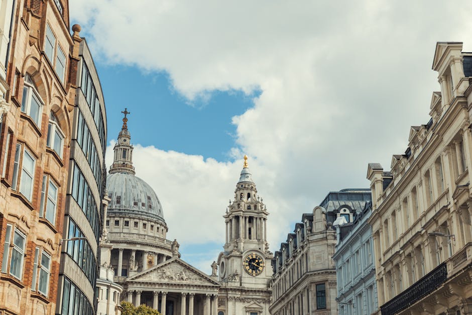 Photograph showing the exterior view of a historic church with a large domed roof and an ornate clock tower, situated between modern glass-fronted office buildings on the left and classic stone façades on the right. Inside the setting, a man from Man with Van St Pauls Cray is engaged in a home relocation process, loading or unloading cardboard boxes and wrapped furniture onto or from a van parked nearby on the pavement. The scene captures the loading process in an urban environment, with clear weather and a partly cloudy sky overhead. The image highlights the logistics of furniture transport and packing during an urgent move, with visible packaging materials such as cardboard boxes and plastic wraps, and the classic architecture contrasting with the modern surroundings, reflecting a typical cityscape where packing and moving services are often required.