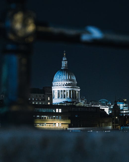 A busy city street scene in London during the evening with traffic including red double-decker buses and cars, leading towards the prominent St. Paul's Cathedral with its large domed roof and classical architecture illuminated against a dark blue sky. The street is lined with a variety of historic and modern buildings, some of which display shop signs and streetlights. In the foreground, pedestrians can be seen walking along the pavement. The image captures the bustling atmosphere typical of central London, providing a contextual backdrop relevant to house removals and moving services offered by Man with Van St Pauls Cray, especially for clients needing efficient logistics within the city.