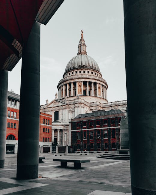 View of St. Paul's Cathedral in the City of London, taken from a low angle between two large cylindrical columns that frame the foreground. The cathedral's iconic domed roof and detailed stone architecture are prominently visible under a cloudy sky. Surrounding the cathedral are modern red-brick and gray buildings, with an open paved area featuring a few benches and steps. The perspective suggests the photo was taken from an outdoor courtyard or public square, highlighting the contrast between the historic cathedral and nearby structures. In the context of house removals or furniture transport, this scene could relate to city-based moving logistics or urban packing and loading processes near central London landmarks, as a subtle nod to the professional services offered by Man with Van St Pauls Cray.