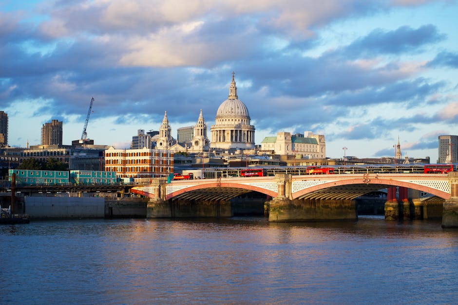 View of St. Paul's Cathedral in the City of London, taken from a low angle between two large cylindrical columns that frame the foreground. The cathedral's iconic domed roof and detailed stone architecture are prominently visible under a cloudy sky. Surrounding the cathedral are modern red-brick and gray buildings, with an open paved area featuring a few benches and steps. The perspective suggests the photo was taken from an outdoor courtyard or public square, highlighting the contrast between the historic cathedral and nearby structures. In the context of house removals or furniture transport, this scene could relate to city-based moving logistics or urban packing and loading processes near central London landmarks, as a subtle nod to the professional services offered by Man with Van St Pauls Cray.