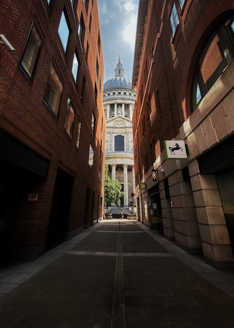 A narrow alleyway between two tall brick buildings leading towards St Paul's Cathedral visible in the background with its dome and classical architectural details. The alley features a paved walkway with a central drain and is lined with side windows and entrance points. On the right side, attached to the building, is a green and white sign with a black horse logo, indicating a business establishment. The scene is set during daytime with partly cloudy skies, and the lighting creates shadowed areas along the building facades, emphasizing the depth and verticality of the surroundings. This urban environment showcases an area where house removals or furniture transport could take place, contextualising [COMPANY_NAME]'s services related to home relocation or moving logistics.