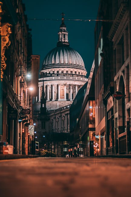 A busy street scene outside a historic building with a large, domed roof and classical architectural features, surrounded by modern red-brick and glass-fronted residential buildings. Pedestrians, dres