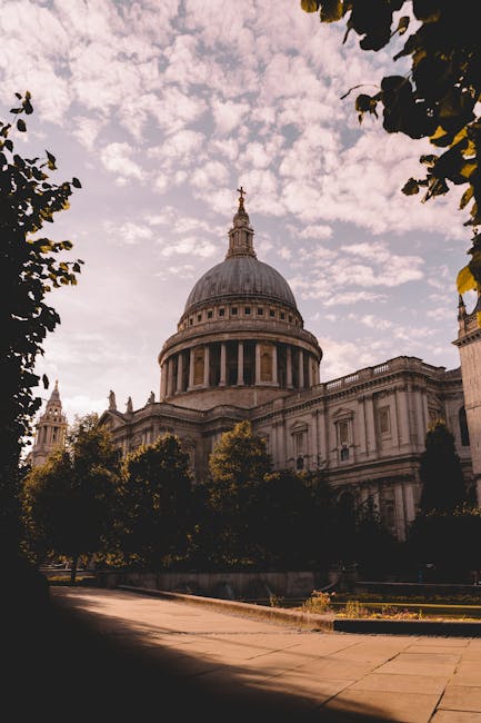 A daytime outdoor scene featuring a statue of a person standing on a pedestal, set against a backdrop of historic buildings with a large domed structure and a spire, likely St Paul's Cathedral in Lond
