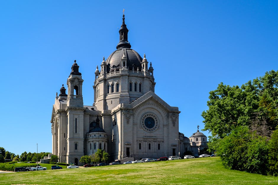 A large, historic church with a prominent dome and elaborate architectural details, situated on a well-maintained grassy area. The church features a tall central spire atop the dome, with smaller decorative towers on either side, and arched windows along its stone facade. Surrounding the church are several parked cars along the street, indicating nearby parking facilities. To the right, a lush green tree partially obscures the view, adding natural contrast to the scene. The sky above is clear and bright blue, suggesting a sunny day. The setting appears to be in a peaceful, suburban area, which may be relevant to house removals or home relocation services such as those provided by Man with Van St Pauls Cray. This location exemplifies typical street-side architecture and outdoor space used during the loading process of furniture transport or packing during a house move, with no visible movement or personnel in the image.