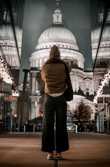 A woman standing in front of a large illuminated dome at night, with her back facing the camera. She is wearing a brown beanie, a tan jacket, and dark wide-leg pants, with a black bag slung over her shoulder. The scene is set on a paved area with string lights and storefronts visible on either side, suggesting a city square or public space. The bright dome architecture is the focal point, lit up against the dark night sky. This setting relates to urban environments where house removals and home relocation services by Man with Van St Pauls Cray can be relevant, especially in contexts of moving within or to city centres or popular districts.